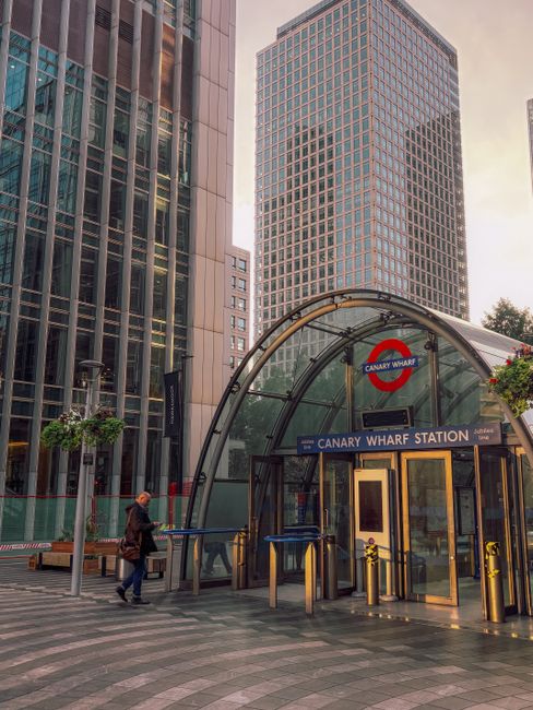 A panoramic view of Canary Wharf's modern skyline featuring tall glass skyscrapers under a partly cloudy sky, with a lush green park in the foreground. The buildings include contemporary office towers