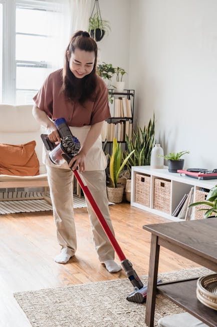A person using a vacuum cleaner with a yellow body and black hose attachment is performing surface cleaning on a patterned area rug within a residential living space. The rug features floral motifs in