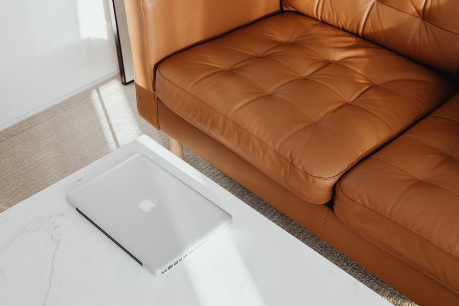 Close-up view of a tan leather sofa with tufted cushions positioned in a well-lit living room, next to a white marble coffee table. On the table, a closed MacBook laptop is placed, with a shadow cast